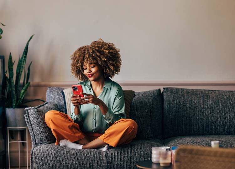 A smiling woman using her smartphone while sitting on the sofa in the living room.