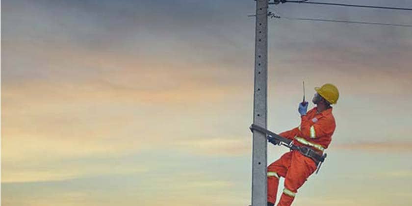 A man in an orange suit and yellow helmet is strapped by a safety belt to an electric pole