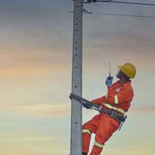 A man in an orange suit and yellow helmet is strapped by a safety belt to an electric pole