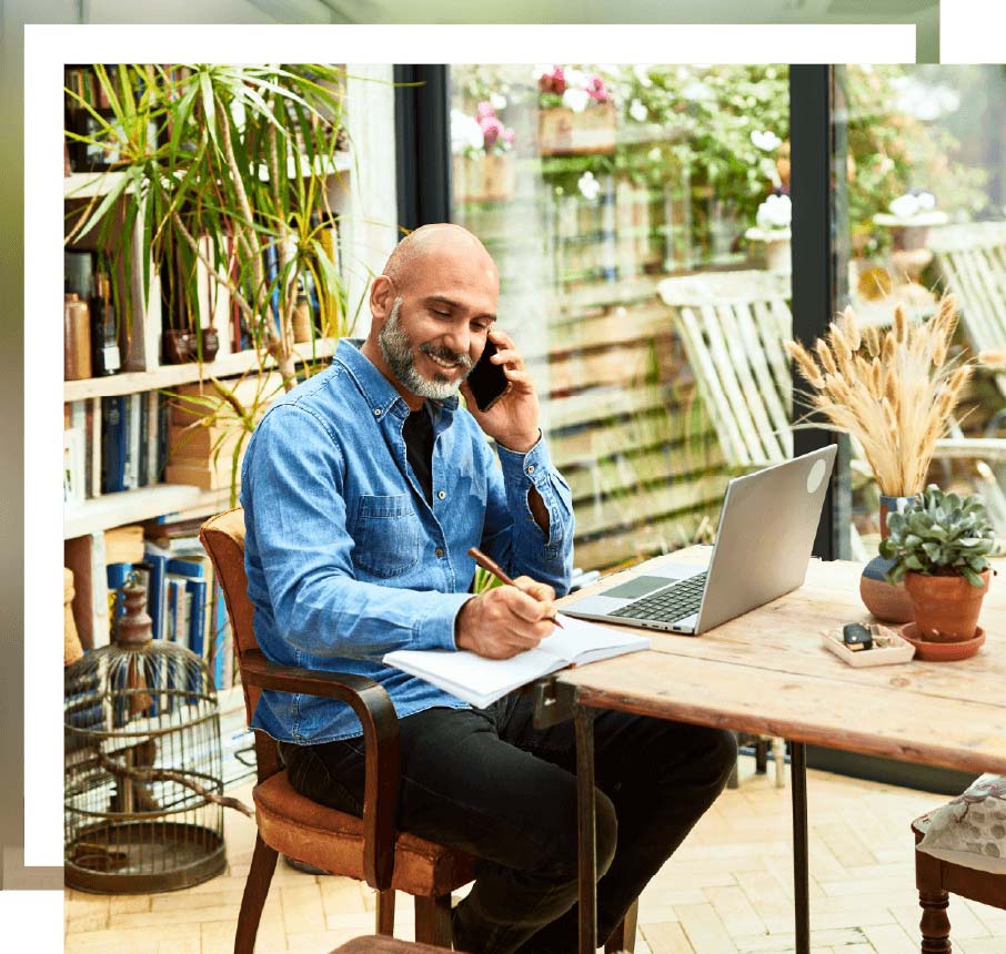 Man on the phone writing on a pad at a table with plants
