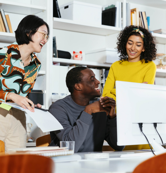 Two women standing and a man sat at a desk, smiling.